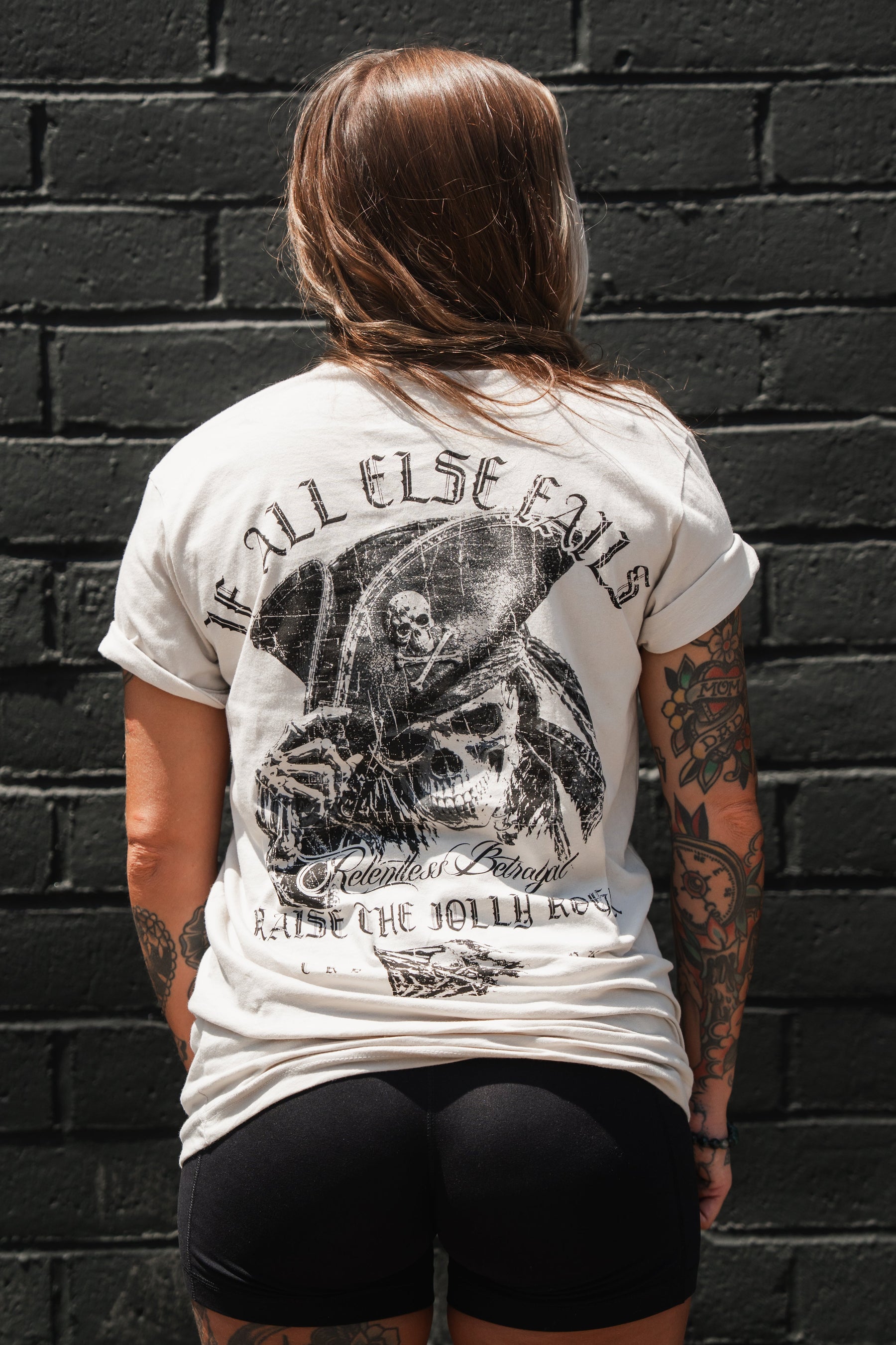 A tattooed individual stands facing a black brick wall, wearing the Relentless Betrayal “Raise the Jolly Roger Bone” T-shirt—featuring bold text and a grim reaper graphic—paired with black shorts for a striking pirate-inspired streetwear look.