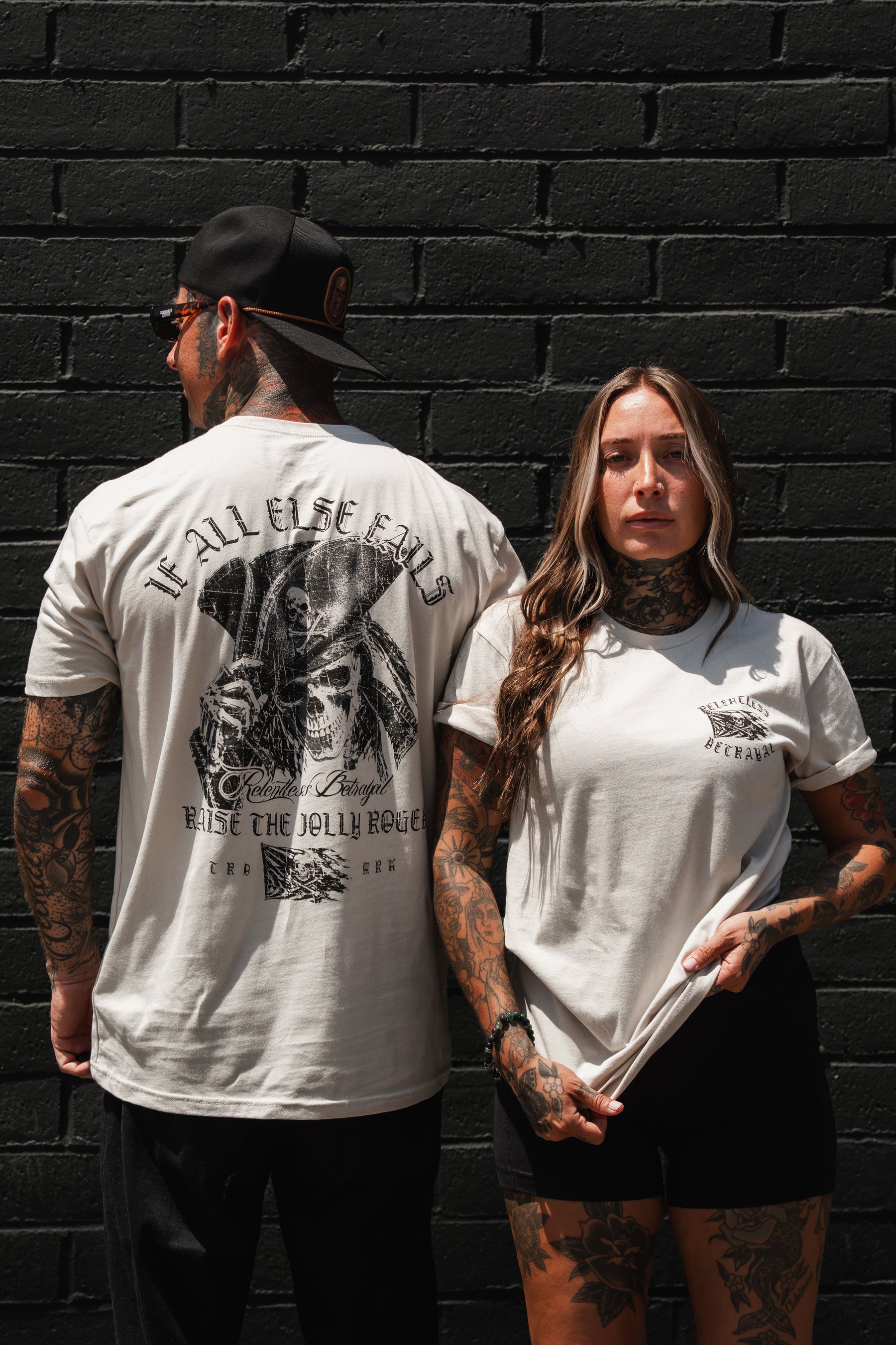 Two tattooed individuals in light-colored t-shirts stand against a black brick wall; one faces forward, while the other turns to showcase the striking graphic on the back of their Relentless Betrayal “Raise the Jolly Roger Bone T-Shirt,” a piece of pirate streetwear boldly featuring distinctive skulls and crossbones.