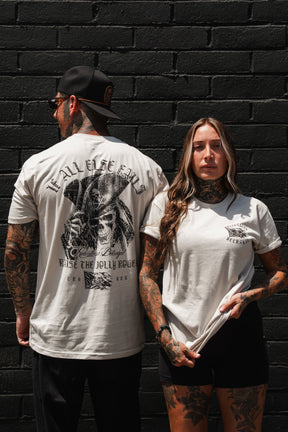 Two tattooed individuals in light-colored t-shirts stand against a black brick wall; one faces forward, while the other turns to showcase the striking graphic on the back of their Relentless Betrayal “Raise the Jolly Roger Bone T-Shirt,” a piece of pirate streetwear boldly featuring distinctive skulls and crossbones.
