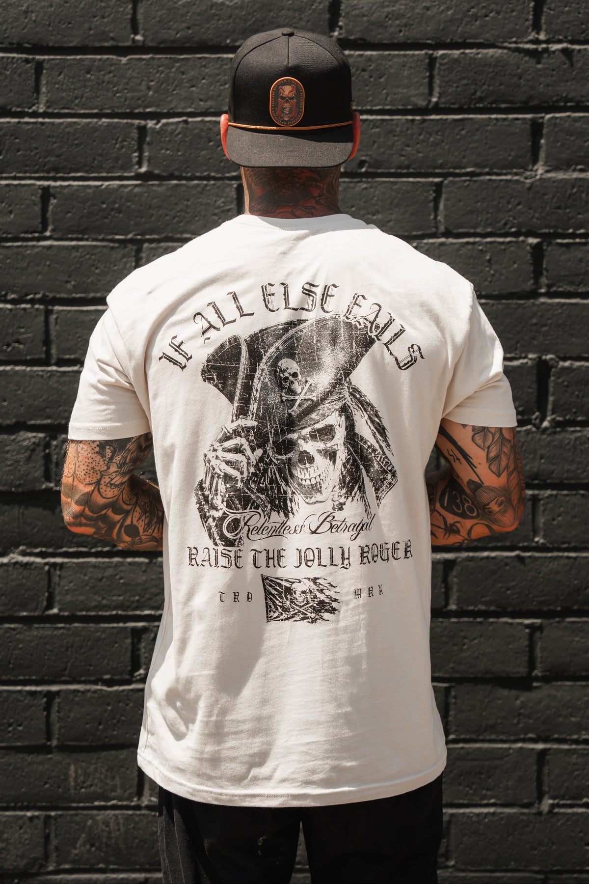 A tattooed individual in a black cap stands facing a black brick wall, wearing the Raise the Jolly Roger Bone T-Shirt by Relentless Betrayal. This bold piece of pirate streetwear showcases a striking pirate skeleton illustration and themed text.