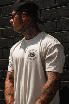 A heavily tattooed man with a rose tattoo on his neck stands against a black brick wall, wearing sunglasses, a backwards cap, and the white “Raise the Jolly Roger Bone” T-shirt by Relentless Betrayal.