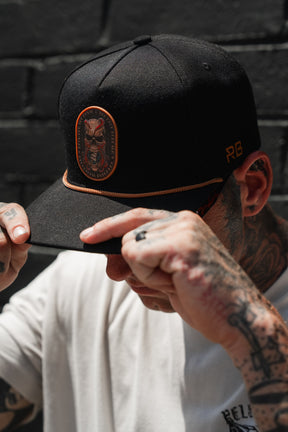 A person adjusts the Relentless Betrayal “Between The Devil & Deep Blue Sea” snapback, featuring a black design with an embroidered patch and orange trim, while wearing a white shirt. Tattoos are visible on their hands and arms against a dark brick wall background.