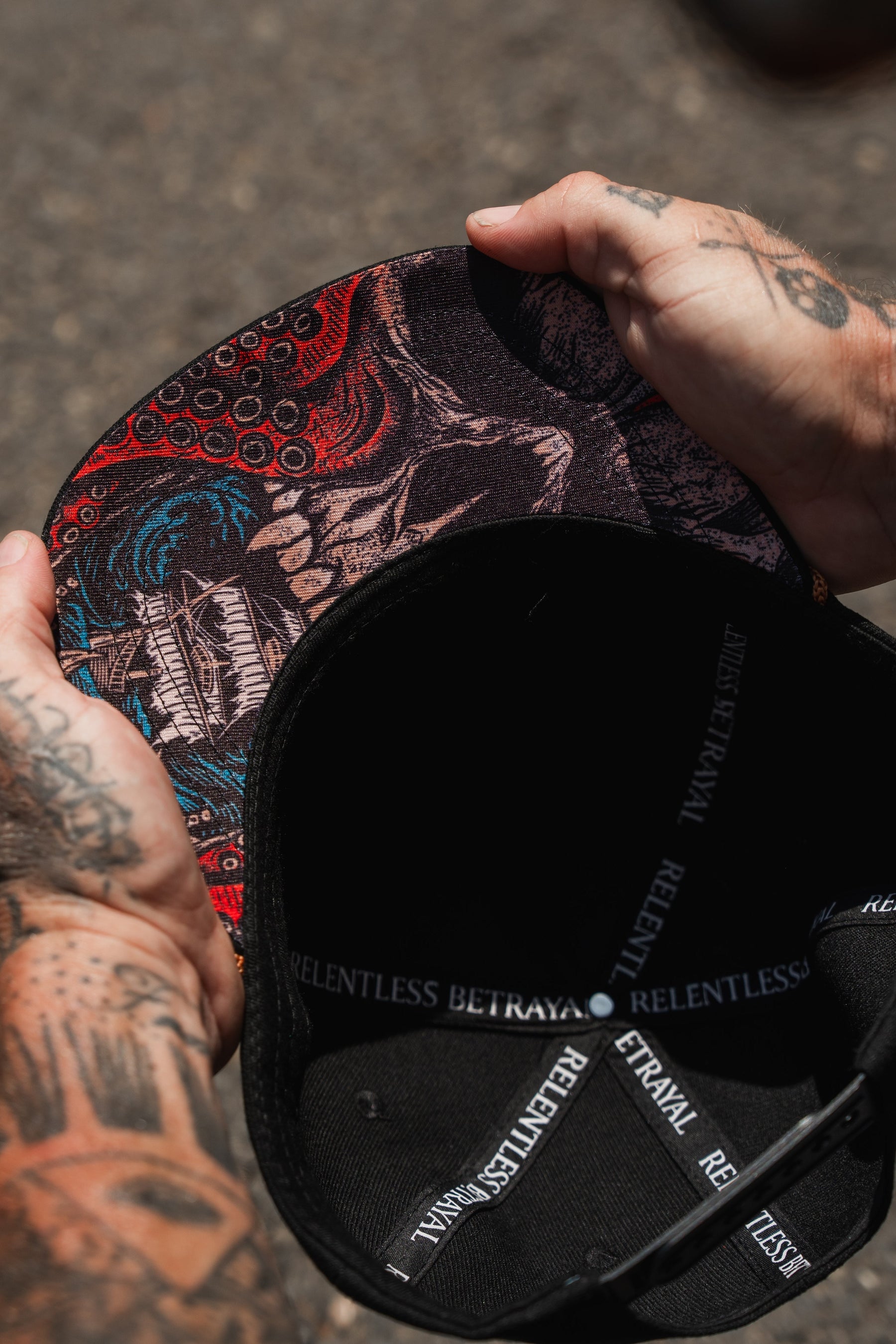A tattooed individual holds an open Between The Devil & Deep Blue Sea Snapback by Relentless Betrayal, showcasing vibrant artwork on the brim and the RELENTLESS BETRAYAL text along the inner lining.