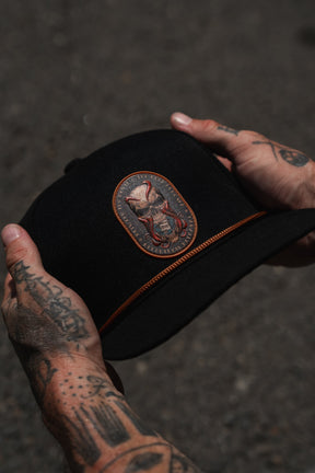 Tattooed hands holding the Relentless Betrayal “Between The Devil & Deep Blue Sea” snapback—a black cap adorned with a brown embroidered patch featuring a stylized design—photographed against a blurred dark background.
