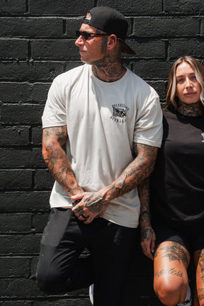 Two tattooed individuals lean against a black brick wall; the man wears a cap, sunglasses, and a white “Raise the Jolly Roger Bone” T-shirt by Relentless Betrayal, while the woman sports a black shirt and shorts.