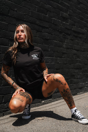 A tattooed individual in the Relentless Betrayal “Raise the Jolly Roger” unisex cotton t-shirt and sneakers squats on a sunlit sidewalk against a black brick wall, gazing to the side.