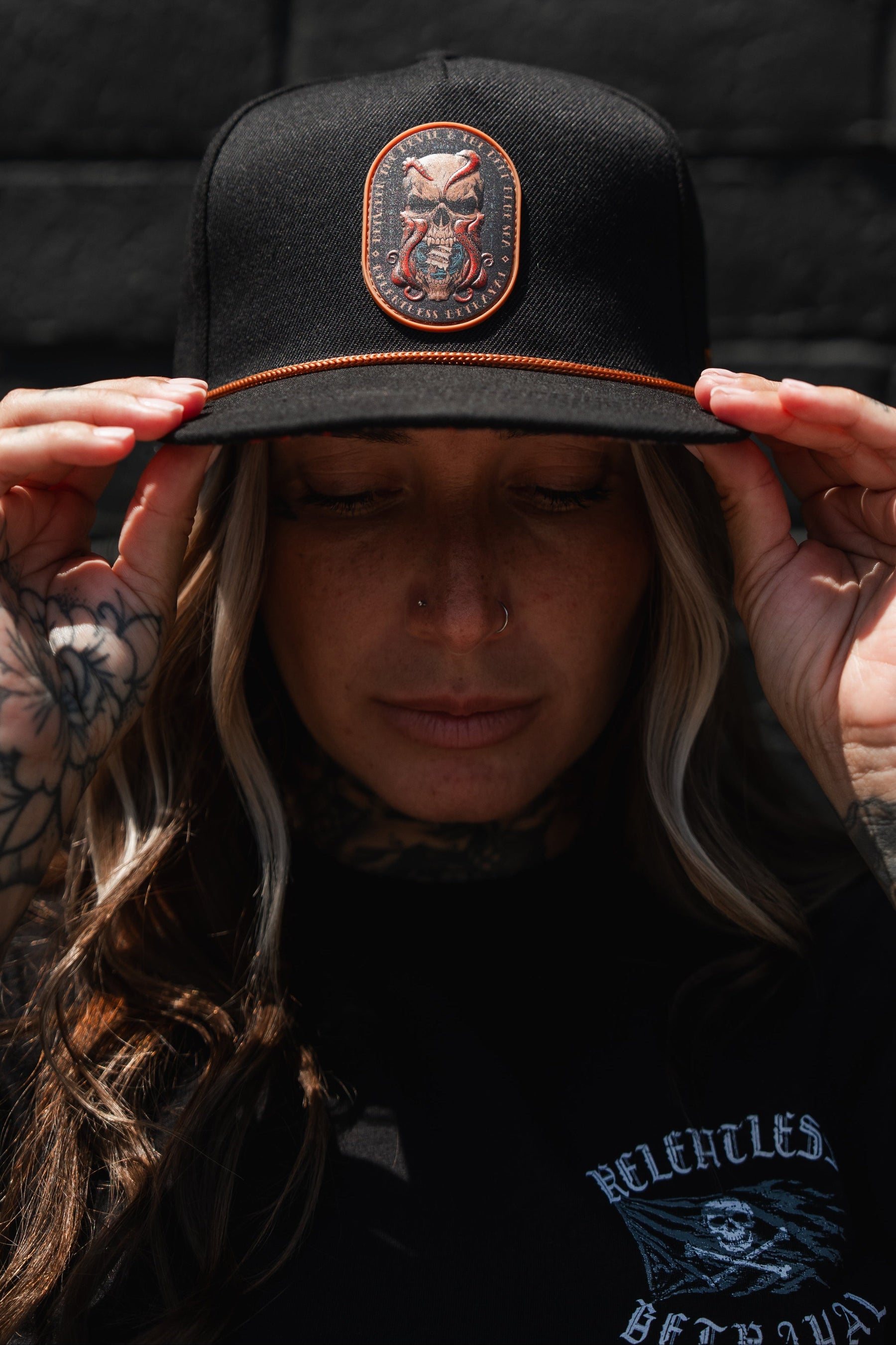 A tattooed individual adjusts the Between The Devil & Deep Blue Sea Snapback by Relentless Betrayal while outdoors. The black hat showcases an embroidered skull patch for a bold, edgy style. They also have a nose piercing and are dressed in a black shirt.