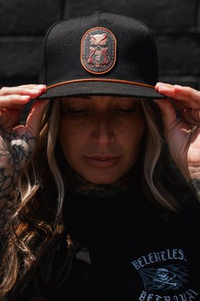 A tattooed individual adjusts the Between The Devil & Deep Blue Sea Snapback by Relentless Betrayal while outdoors. The black hat showcases an embroidered skull patch for a bold, edgy style. They also have a nose piercing and are dressed in a black shirt.