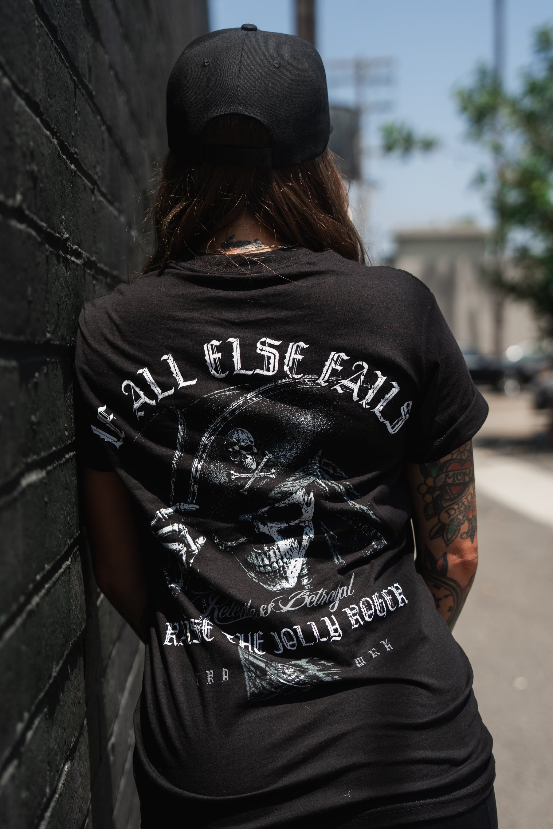 A tattooed person in a black cap stands against a brick wall, wearing the Relentless Betrayal “Raise the Jolly Roger” unisex cotton t-shirt featuring a large skull graphic and “Call Elsefalls” text on the back.