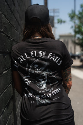 A tattooed person in a black cap stands against a brick wall, wearing the Relentless Betrayal “Raise the Jolly Roger” unisex cotton t-shirt featuring a large skull graphic and “Call Elsefalls” text on the back.