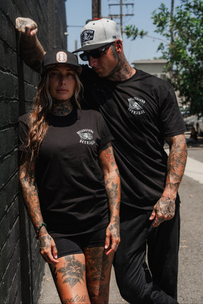 Two tattooed people wearing black “Raise the Jolly Roger” T-Shirts by Relentless Betrayal, along with shorts and hats, stand next to a brick wall outdoors on a sunny day.