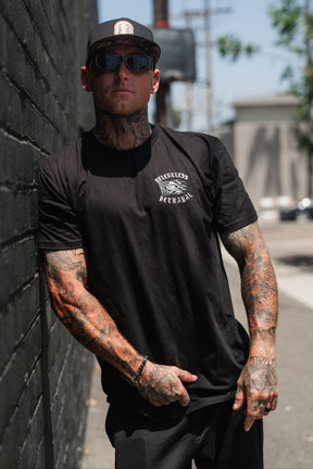 A man with tattoos, sporting sunglasses and a black cap, stands against a brick wall outdoors on a sunny day while wearing the Relentless Betrayal “Raise the Jolly Roger” T-shirt.