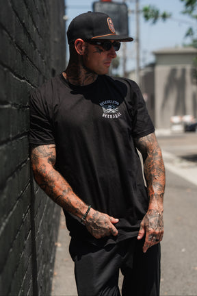 A tattooed man sporting sunglasses and a black cap leans against a black brick wall outdoors, wearing the “Raise the Jolly Roger T-Shirt” by Relentless Betrayal paired with black pants.