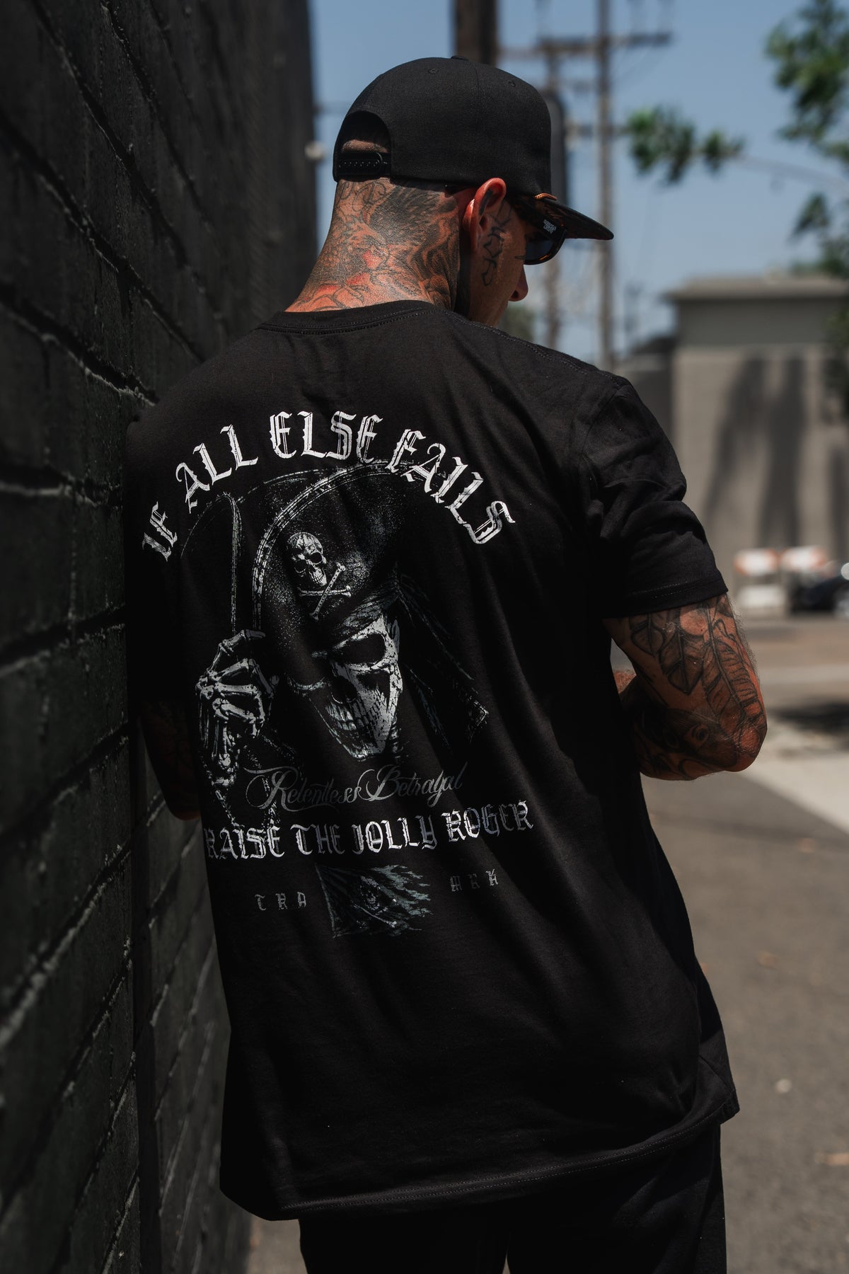 A tattooed individual in a black cap and black shirt stands beside a brick wall. The unisex Raise the Jolly Roger T-Shirt by Relentless Betrayal features a striking grim reaper graphic with the phrase “IF ALL ELSE FAILS, RAISE THE JOLLY ROGER,” making it the perfect bold, nautical-themed graphic tee for those seeking standout style.