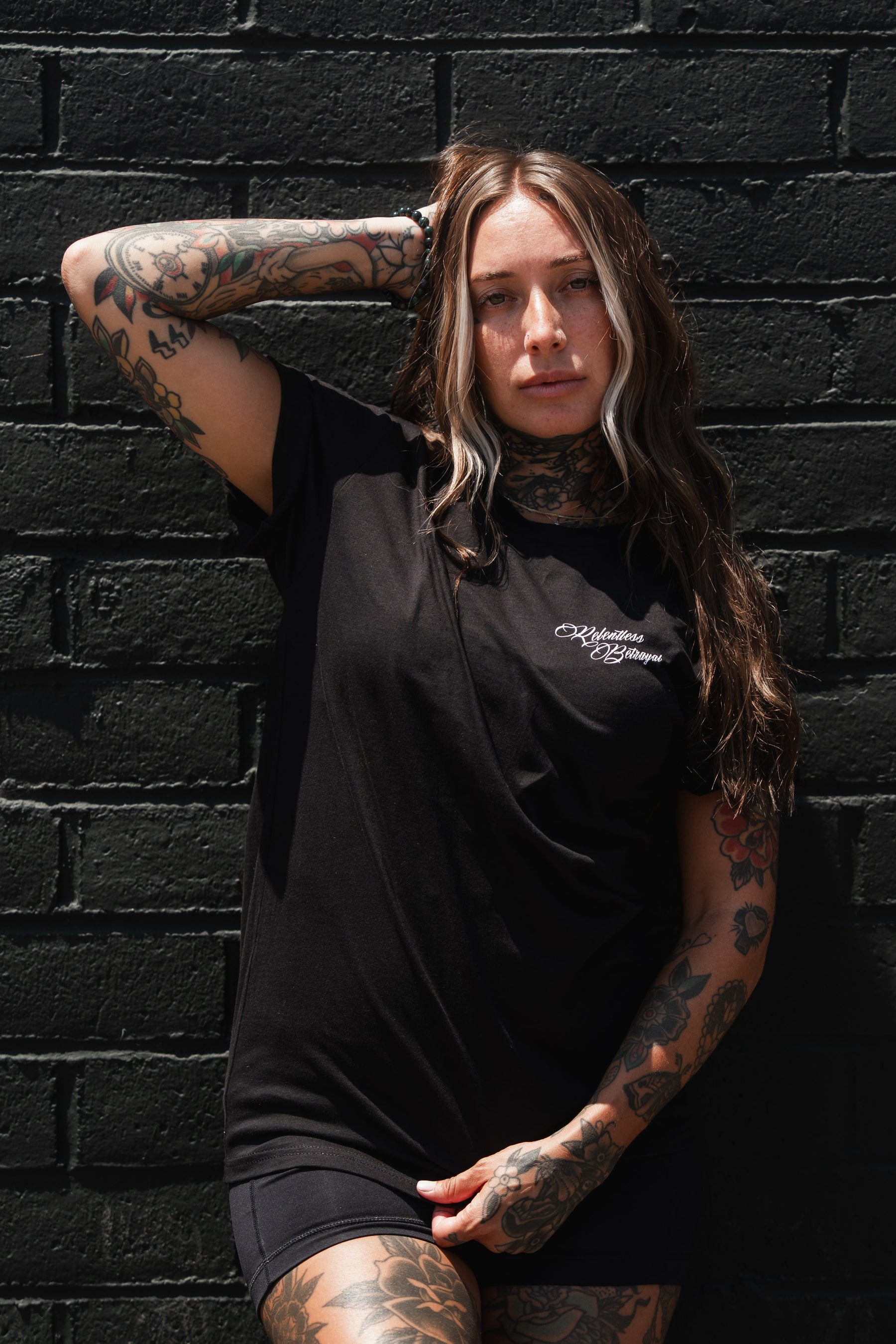A woman with long brown hair and visible tattoos poses against a black brick wall, wearing the Relentless Betrayal Raise the Jolly Roger T-Shirt and black shorts, one arm resting on her head—a bold statement of pirate streetwear style.