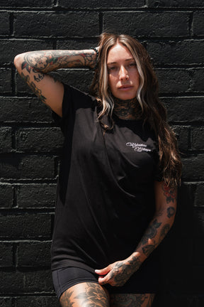 A woman with long brown hair and visible tattoos poses against a black brick wall, wearing the Relentless Betrayal Raise the Jolly Roger T-Shirt and black shorts, one arm resting on her head—a bold statement of pirate streetwear style.