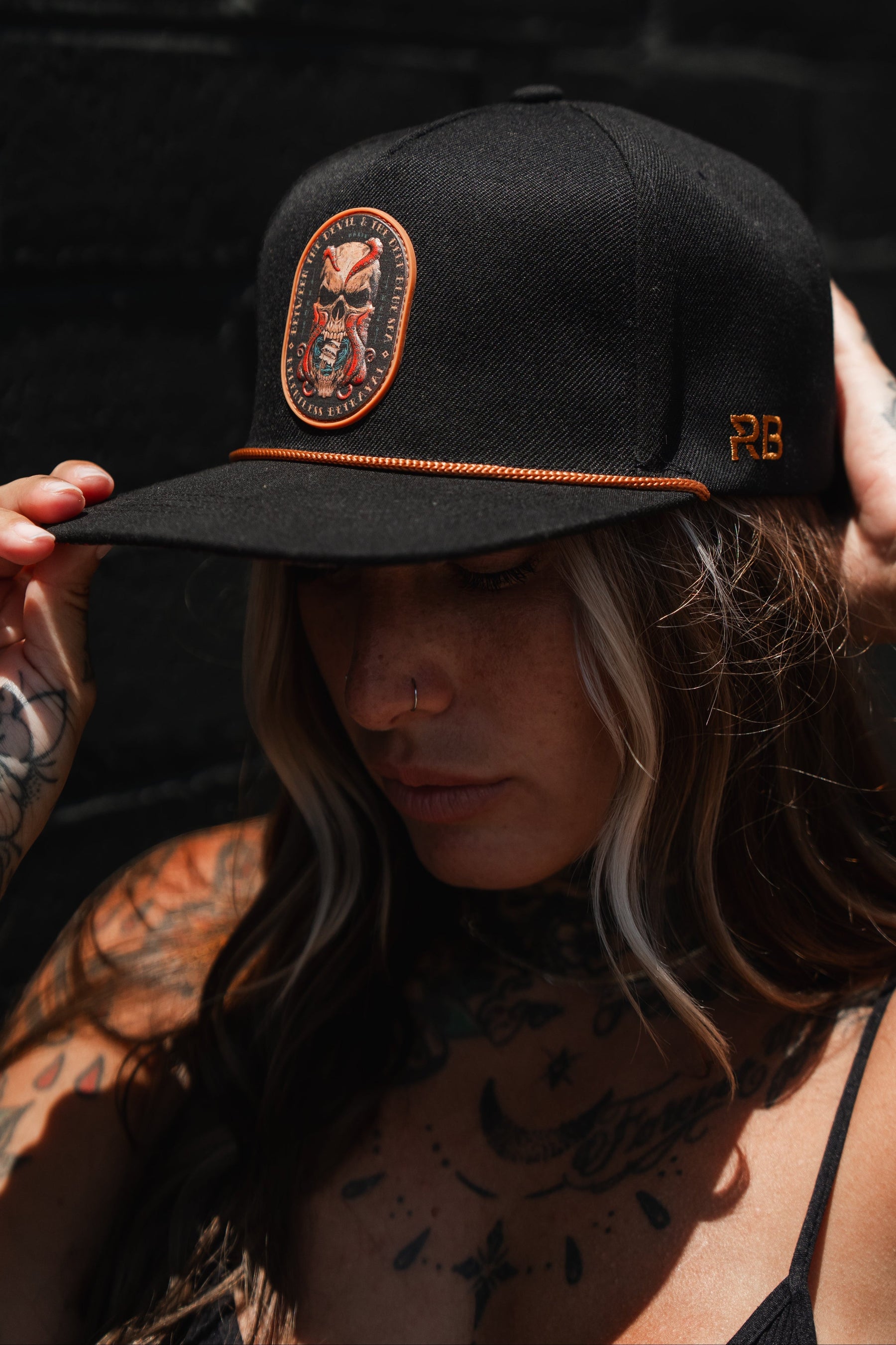 A person with tattoos and a nose ring adjusts the brim of their Relentless Betrayal Between The Devil & Deep Blue Sea Snapback, featuring an embroidered patch and orange trim, using both hands.