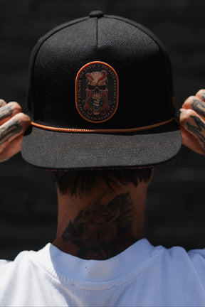 A person, seen from behind, adjusts the Relentless Betrayal “Between The Devil & Deep Blue Sea” Snapback—a black nautical cap featuring a decorative patch. Tattoos are visible on their hands and neck as they wear a white shirt against a dark background.