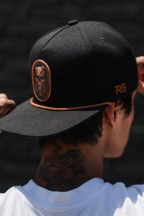A person is seen from behind wearing the Relentless Betrayal “Between The Devil & Deep Blue Sea” snapback, which features a black design with an orange emblem and RB initials, along with a white shirt and a neck tattoo.