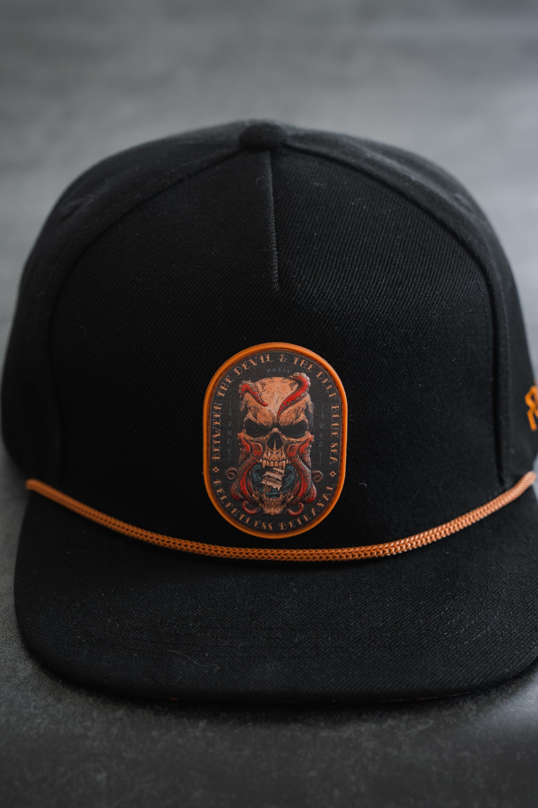 The Between The Devil & Deep Blue Sea Snapback by Relentless Betrayal is a black hat with an orange cord and a front patch featuring a skull with red highlights and surrounding text—perfect for channeling the pirate lifestyle.