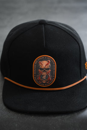 The Between The Devil & Deep Blue Sea Snapback by Relentless Betrayal is a black hat with an orange cord and a front patch featuring a skull with red highlights and surrounding text—perfect for channeling the pirate lifestyle.