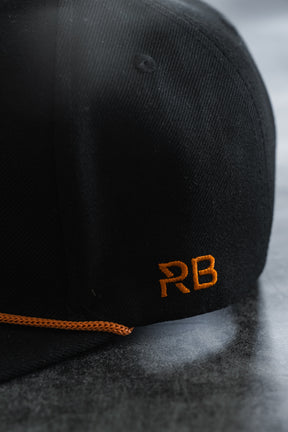 Black cap with orange RB embroidery on the side and an orange trim along the brim, shown on a gray surface—a perfect Between The Devil & Deep Blue Sea Snapback by Relentless Betrayal for those who embrace the pirate lifestyle.