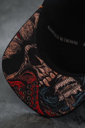 A close-up of the brim of the Relentless Betrayal “Between The Devil & Deep Blue Sea Snapback,” showcasing detailed artwork of a skull, tentacles, and a sailing ship on a dark background.