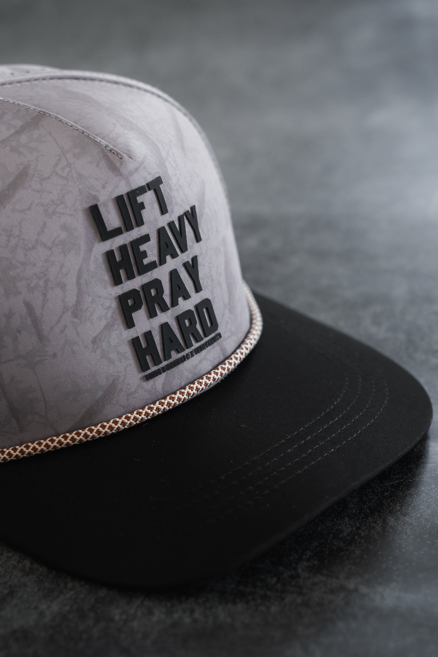 The Lift Heavy. Pray Hard. Deadwood Camo Hydro Snapback Hat by Tango Charlie Apparel features a gray and black design with bold black lettering across the front and a gold braid above the brim, shown here resting on a gray surface. This stylish cap is perfect for those seeking motivation wherever they go.