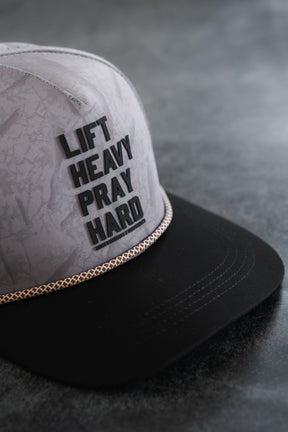 The Lift Heavy. Pray Hard. Deadwood Camo Hydro Snapback Hat by Tango Charlie Apparel features a gray and black design with bold black lettering across the front and a gold braid above the brim, shown here resting on a gray surface. This stylish cap is perfect for those seeking motivation wherever they go.