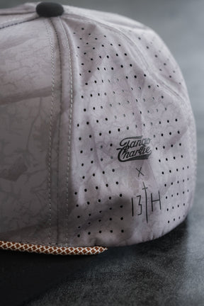 Close-up of the Lift Heavy. Pray Hard. Deadwood Camo Hydro Snapback Hat by Tango Charlie Apparel, featuring a light gray perforated, water-resistant snapback design with a patterned brown and white edge on the brim.