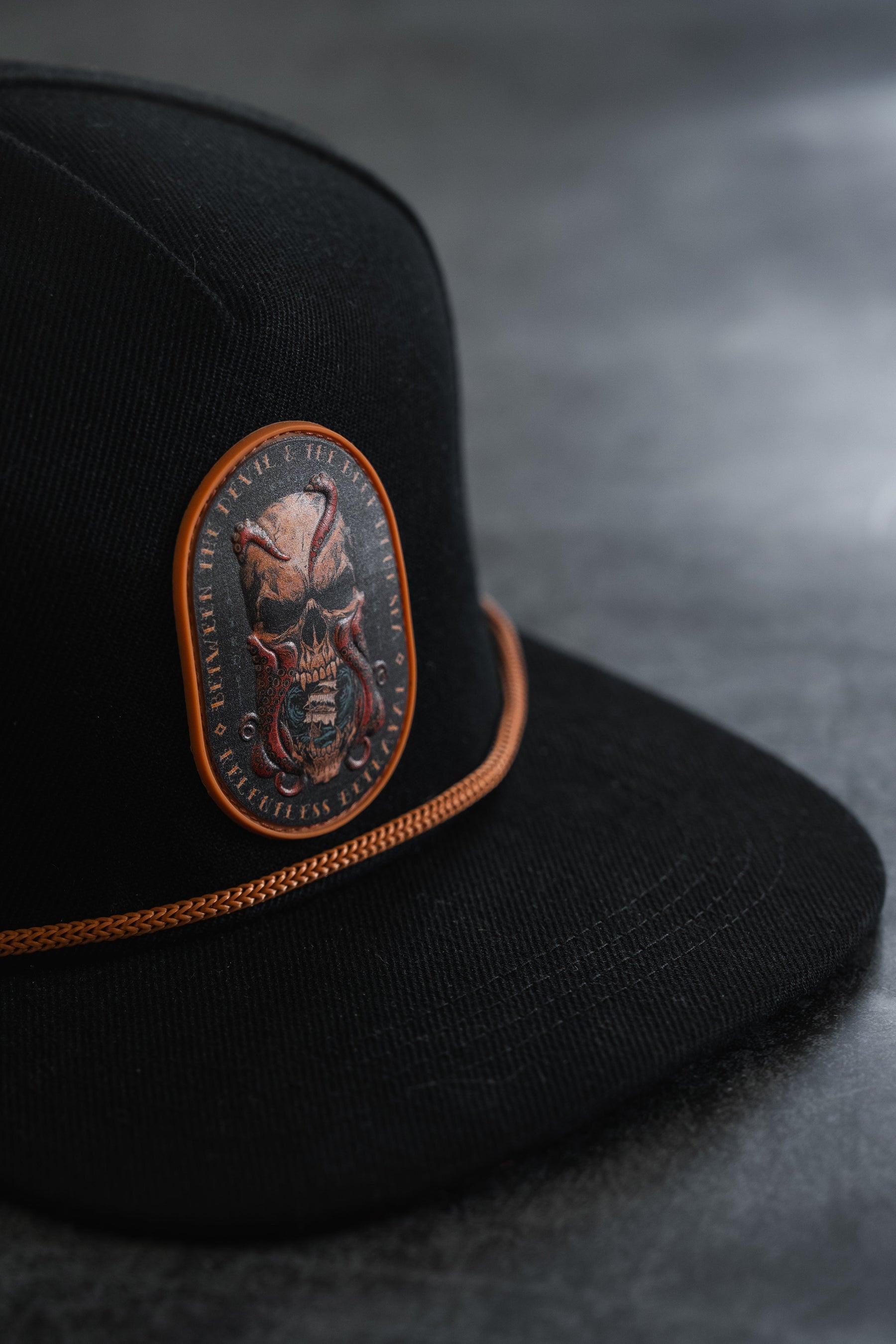 Between The Devil & Deep Blue Sea Snapback by Relentless Betrayal—a black cap featuring an orange-trimmed patch with a fierce skull and tentacles, set against a dark, textured background; this bold nautical snapback makes a striking statement in pirate-inspired style.
