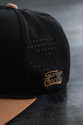Close-up of the Tango Charlie Apparel “Lift Heavy. Trust God. Hydro - Black Snapback Hat,” a black and tan gym hat featuring sweat-resistant fabric, ventilation holes, and the Tango Charlie logo printed on the side panel—combining functional performance and Christian style for those who lift heavy and trust God.