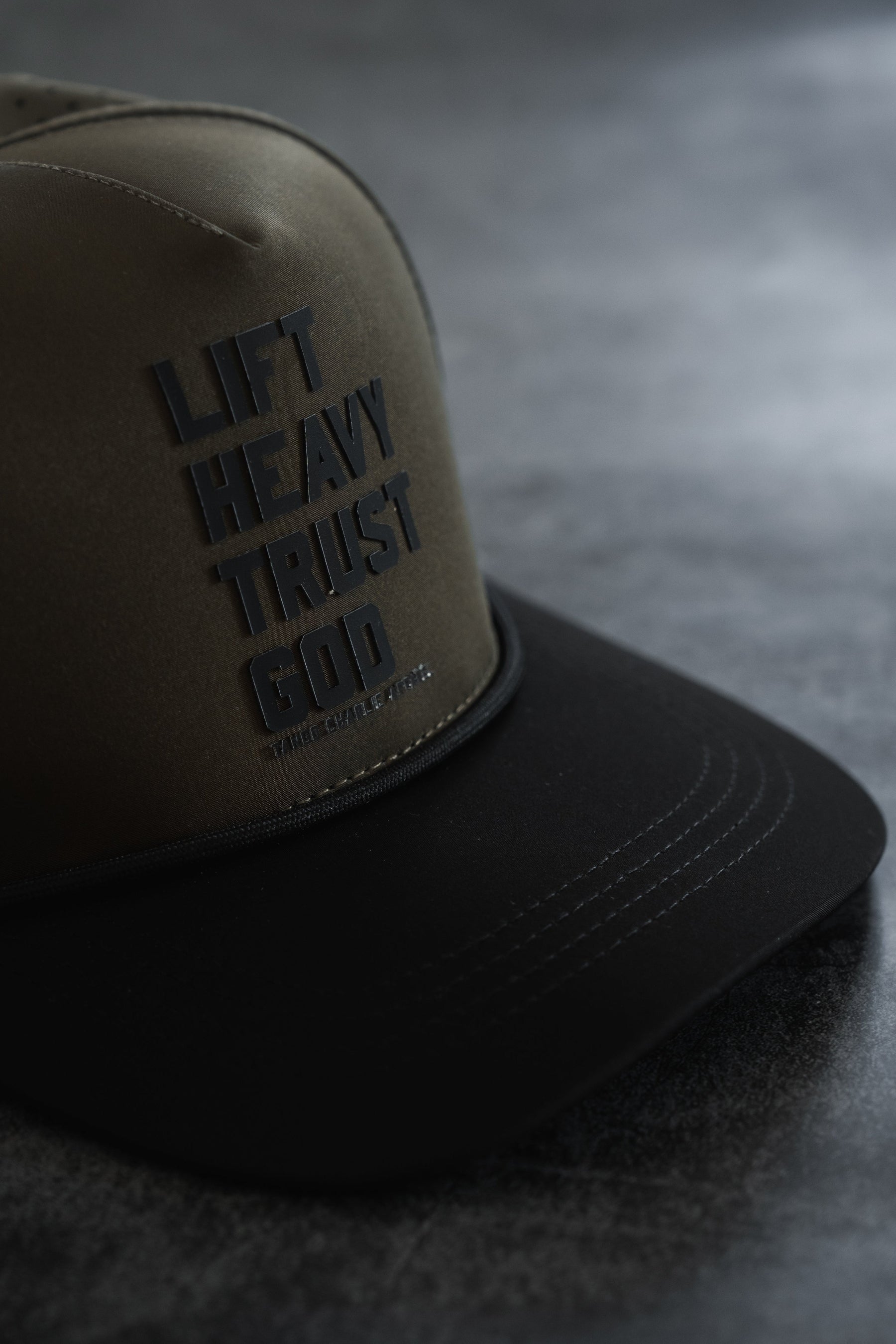 The Lift Heavy. Trust God. Hydro - OD Green Snapback Hat by Tango Charlie Apparel features bold black “LIFT HEAVY TRUST GOD” text on the front panel and is shown resting on a dark surface. Designed to be sweat resistant, this Christian gym hat is ideal for workouts and everyday use, keeping you comfortable while making a powerful statement of faith.