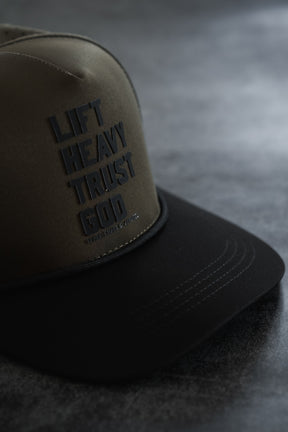 The Lift Heavy. Trust God. Hydro - OD Green Snapback Hat by Tango Charlie Apparel features bold black “LIFT HEAVY TRUST GOD” text on the front panel and is shown resting on a dark surface. Designed to be sweat resistant, this Christian gym hat is ideal for workouts and everyday use, keeping you comfortable while making a powerful statement of faith.