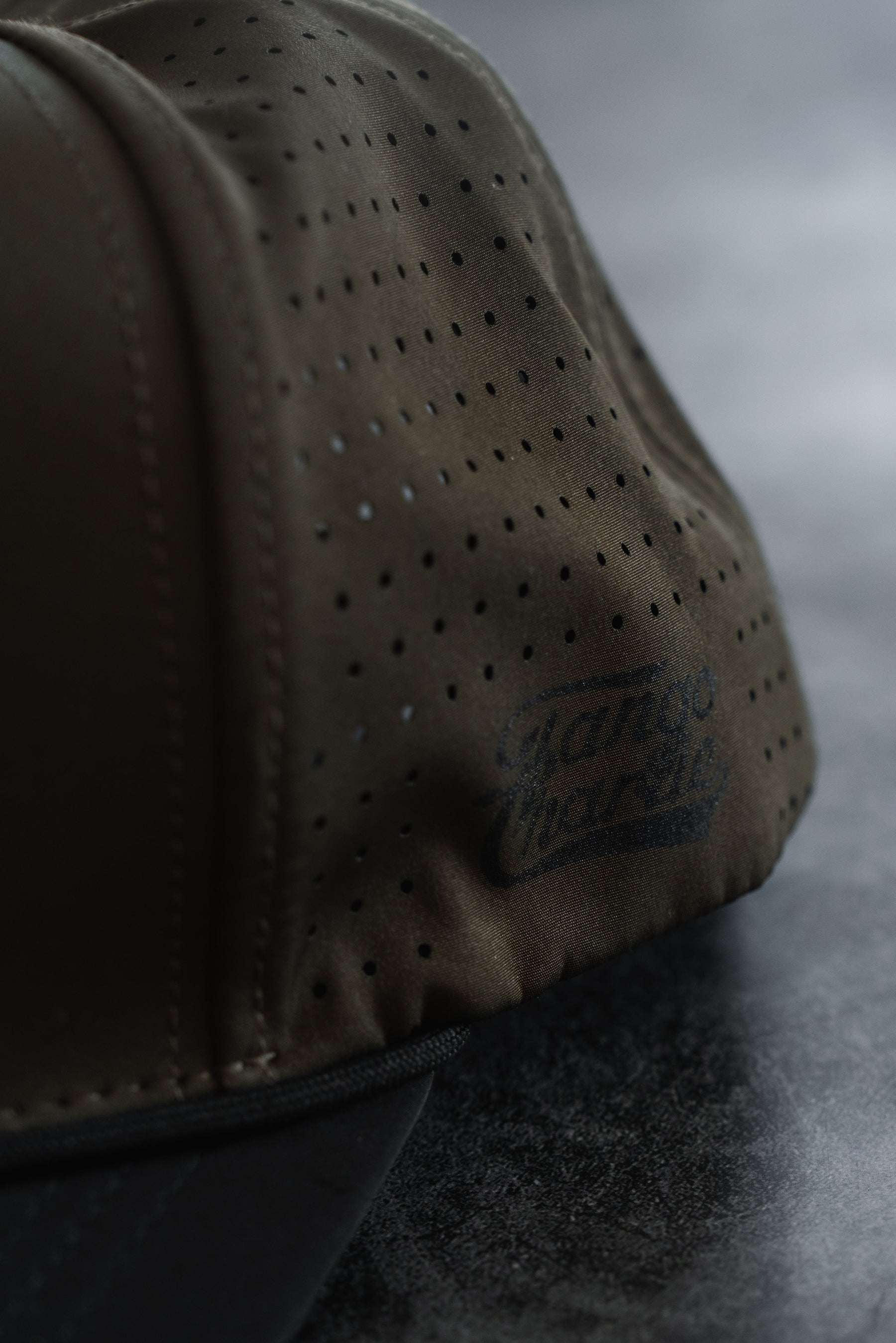 Close-up of an olive green Lift Heavy. Trust God. Hydro - OD Green Snapback Hat by Tango Charlie Apparel, featuring subtle “Tango Charlie” branding on the side and resting on a dark, textured surface. This perforated snapback is sweat-resistant and built for both comfort and durability.
