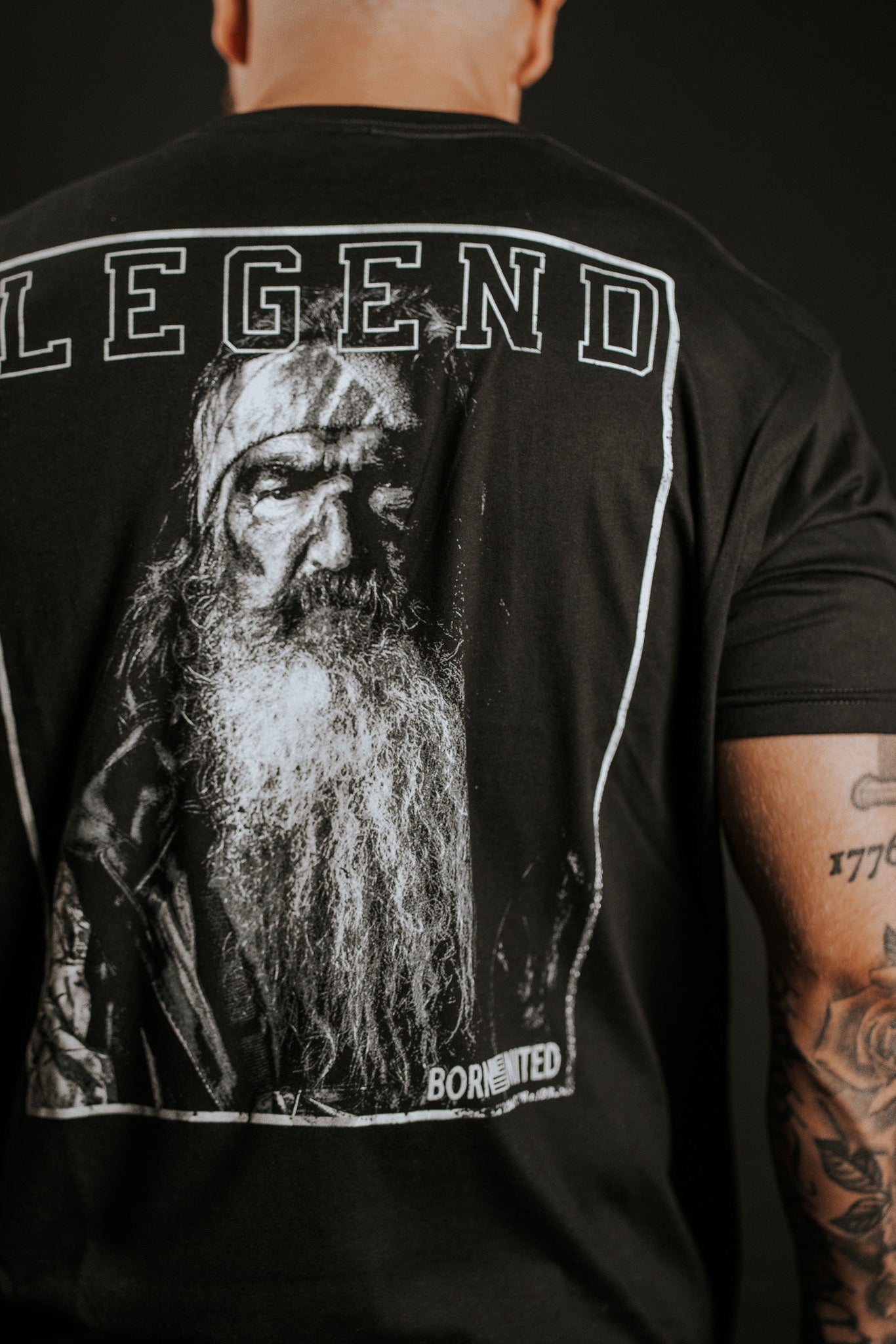 A person with tattooed arms wears the Legend Dynasty T-shirt by Born United Private Label, showcasing a large grayscale portrait of an older man with a long beard and LEGEND printed above the image.