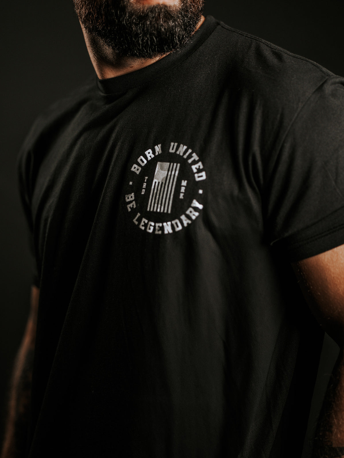 A bearded man wears a black Legend Dynasty t-shirt by Born United Private Label, featuring a white chest emblem with BORN UNITED and BE LEGENDARY encircling a stylized flag. The background is dark.