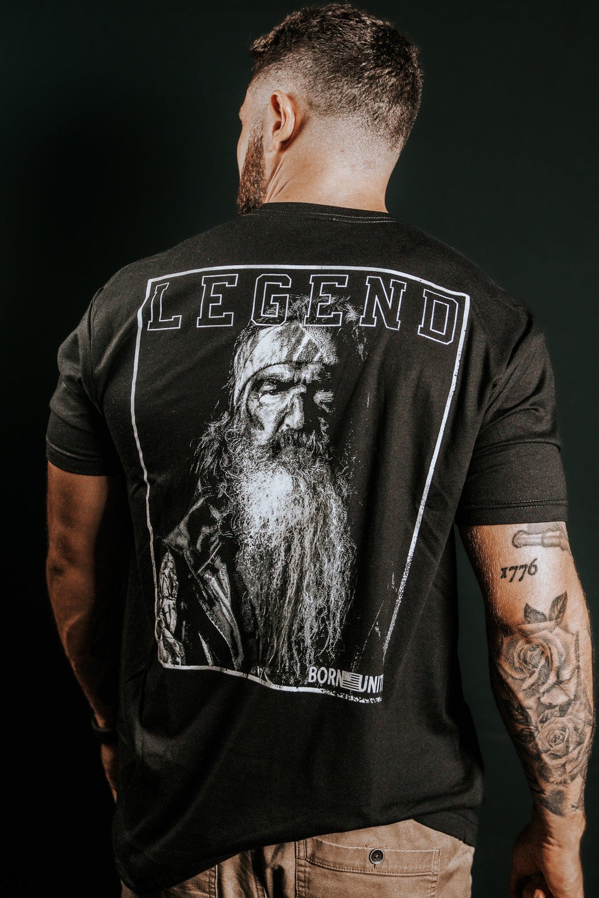 A man with tattoos on his left arm faces away from the camera, wearing the Born United Private Label Legend Dynasty black T-shirt featuring an older bearded mans graphic and LEGEND above the image.