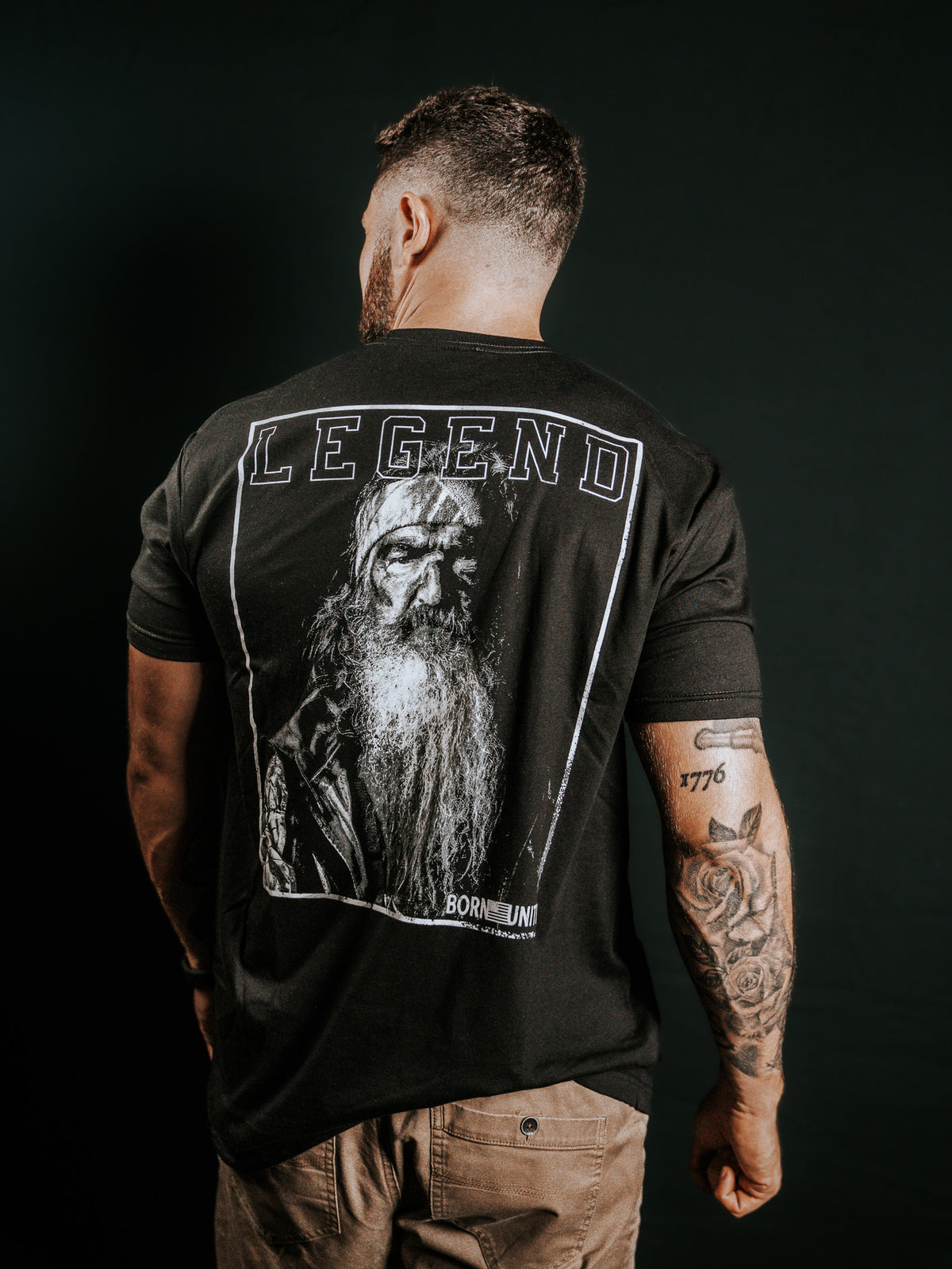 A man with tattoos on his left arm faces away from the camera, wearing the Born United Private Label Legend Dynasty black T-shirt featuring an older bearded mans graphic and LEGEND above the image.
