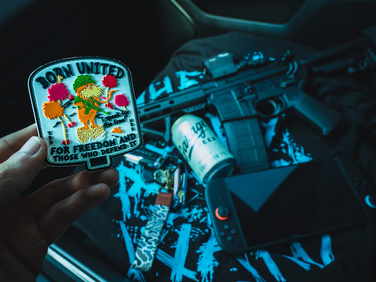 A hand holds the Speak for the Free PVC Patch by Born United Private Label, featuring a Velcro backing, in front of a tabletop with tactical gear, a rifle, an energy drink, keys, and a handheld gaming console.
