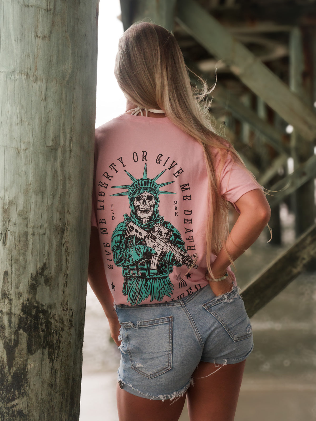 A woman with long blonde hair, wearing Born United Private Labels Liberty Or Death- Heather Pink tee and distressed denim shorts, stands under a pier facing away from the camera.