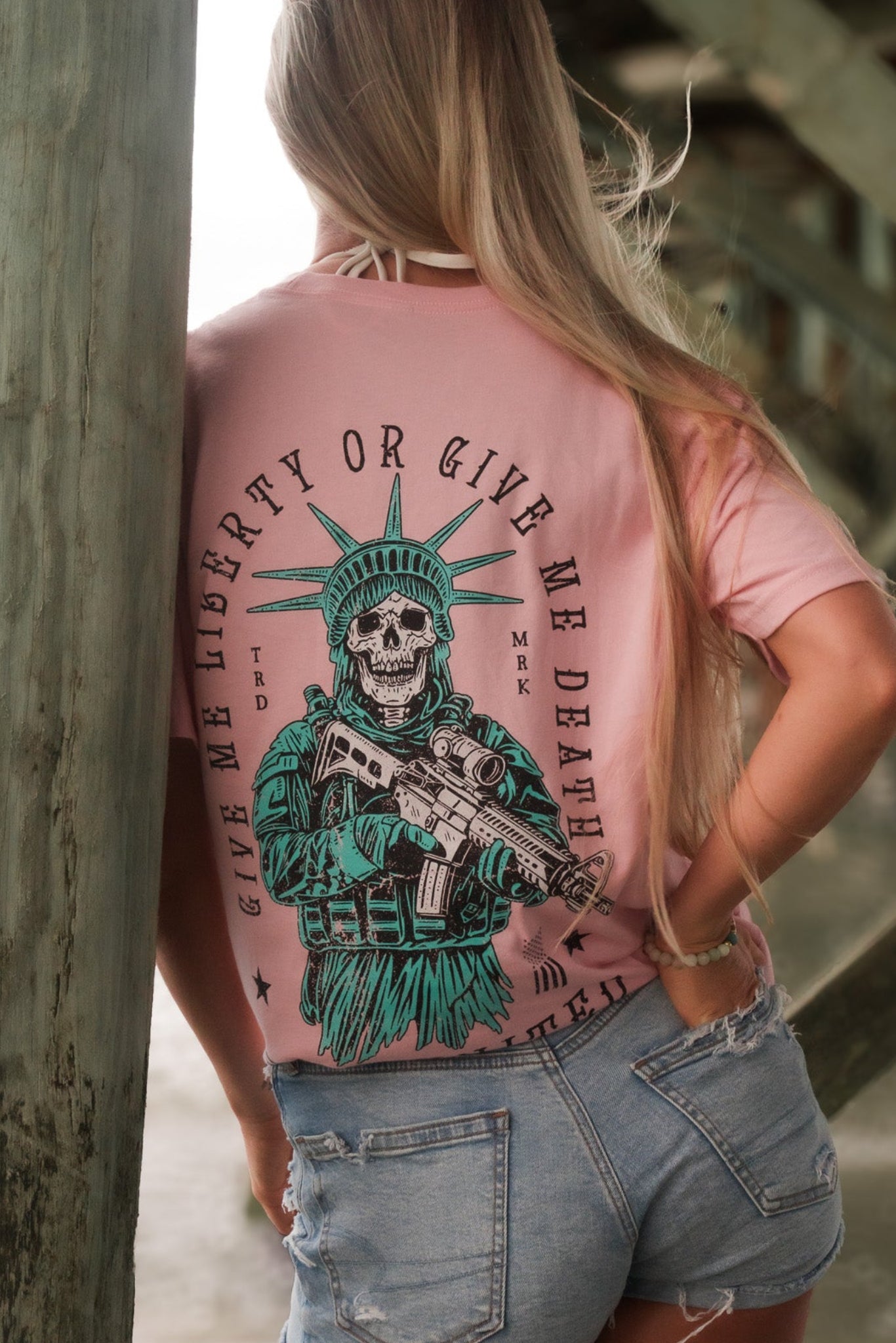 A woman with long blonde hair, wearing Born United Private Labels Liberty Or Death- Heather Pink tee and distressed denim shorts, stands under a pier facing away from the camera.