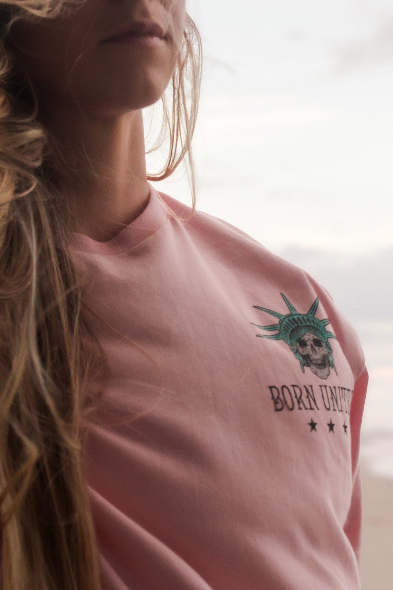 A woman with long blonde hair wears the Born United Private Label Liberty Or Death- Heather Pink tee featuring the Statue of Libertys head and BORN UNIQUE text with stars, standing on a beach at sunset.