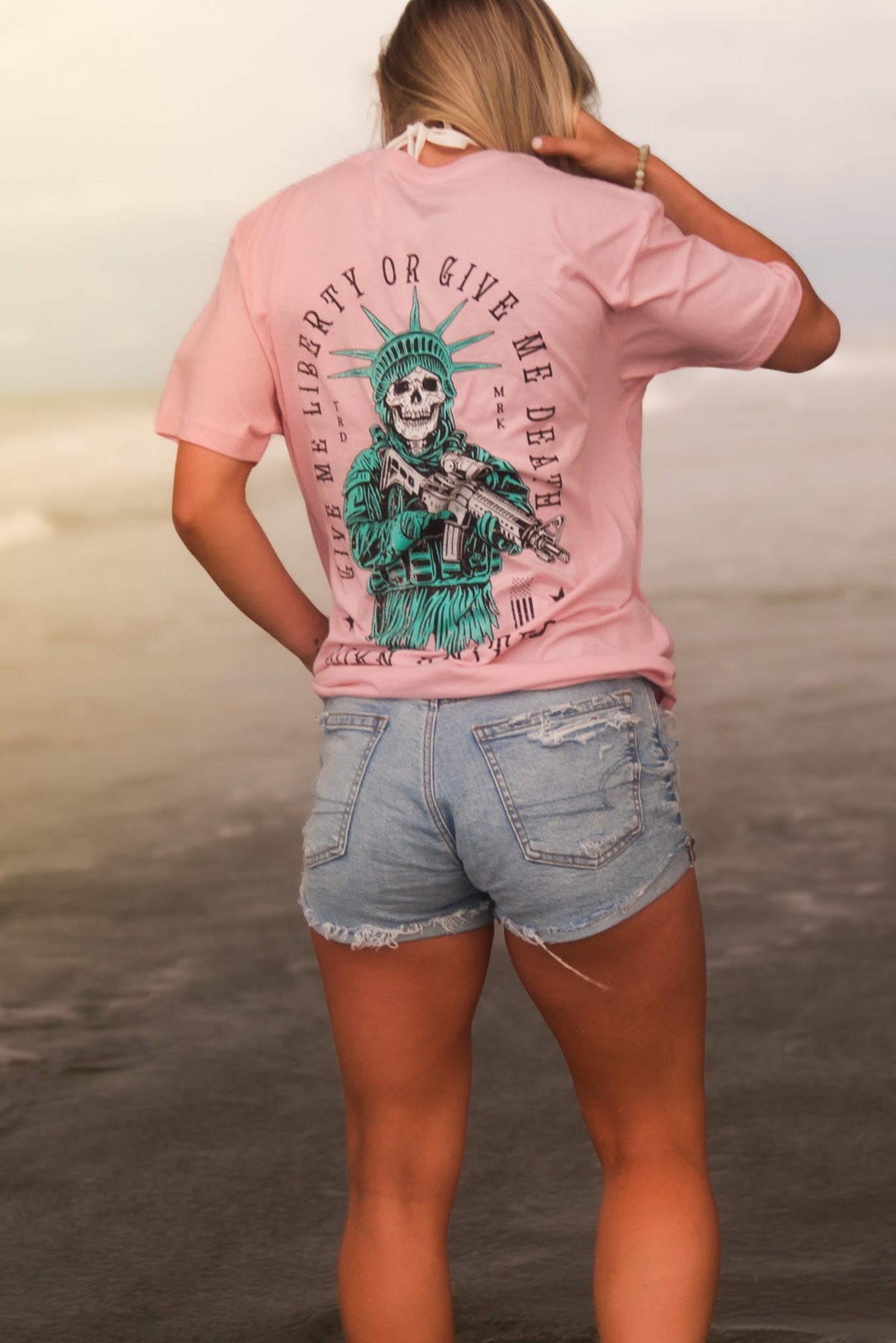 A woman with blonde hair, wearing the Born United Private Label Liberty Or Death- Heather Pink tee, stands in shallow beach water facing away from the camera, paired with denim shorts.