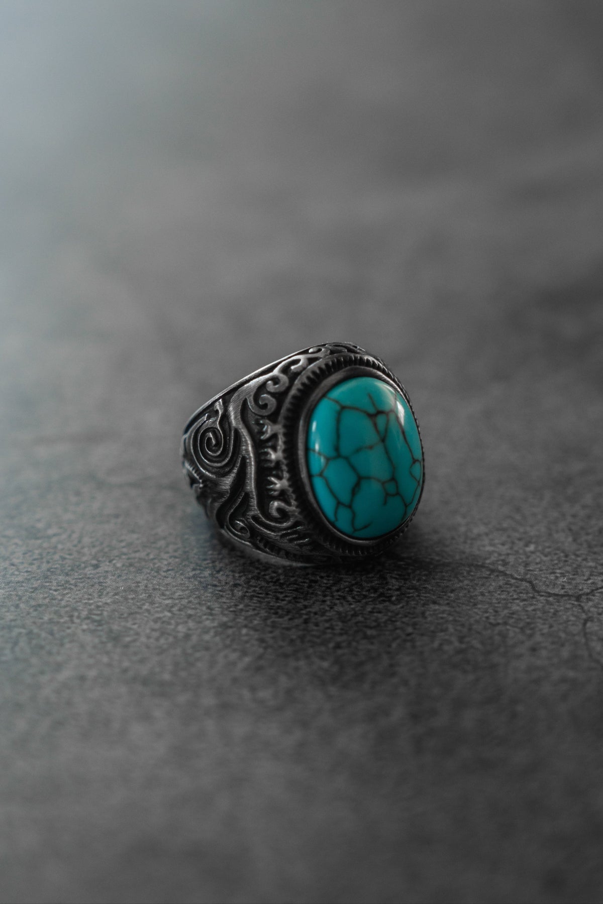 The Into the Storm Ring by Relentless Betrayal showcases a large oval turquoise stone with black veining and ornate silver designs, beautifully displayed on a smooth, gray, stone-like surface.
