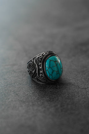 The Into the Storm Ring by Relentless Betrayal showcases a large oval turquoise stone with black veining and ornate silver designs, beautifully displayed on a smooth, gray, stone-like surface.