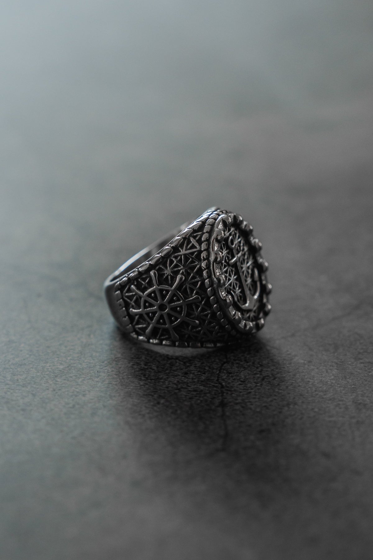The Anchored in Glory Ring by Relentless Betrayal, featuring detailed geometric and circular designs, is displayed on a smooth gray surface for a striking touch of nautical jewelry charm.