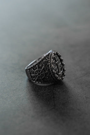 The Anchored in Glory Ring by Relentless Betrayal, featuring detailed geometric and circular designs, is displayed on a smooth gray surface for a striking touch of nautical jewelry charm.