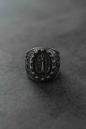 A close-up of the Anchored in Glory Ring by Relentless Betrayal—an ornate, dark stainless steel piece with an engraved anchor symbol, displayed on a gray-black background—ideal for fans of nautical jewelry.