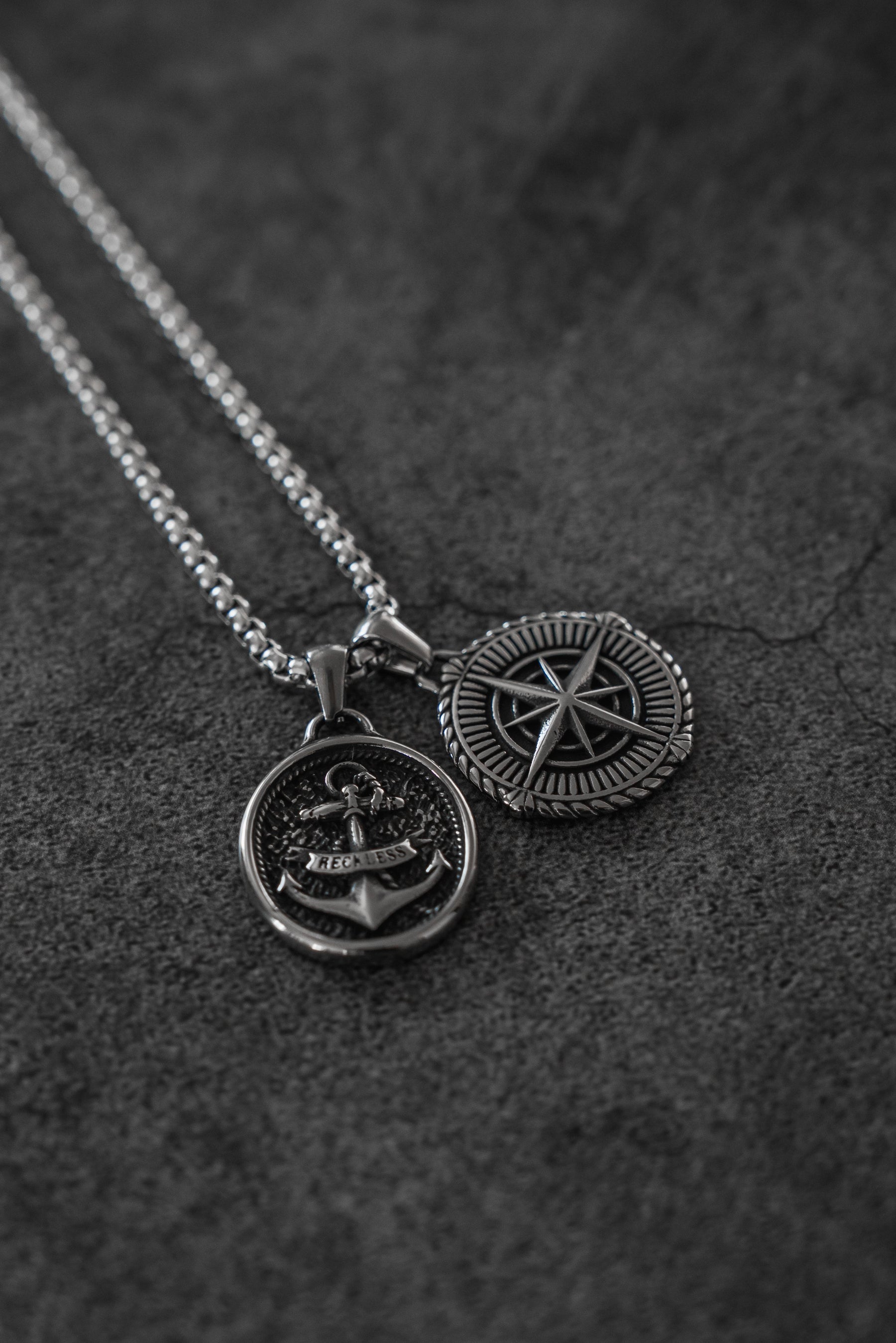 Two Relentless Betrayal Reckless Soul Necklaces with round stainless steel pendants—one with an anchor, one with a compass rose—rest on a dark textured background; each features a 26-inch chain and handcrafted details.